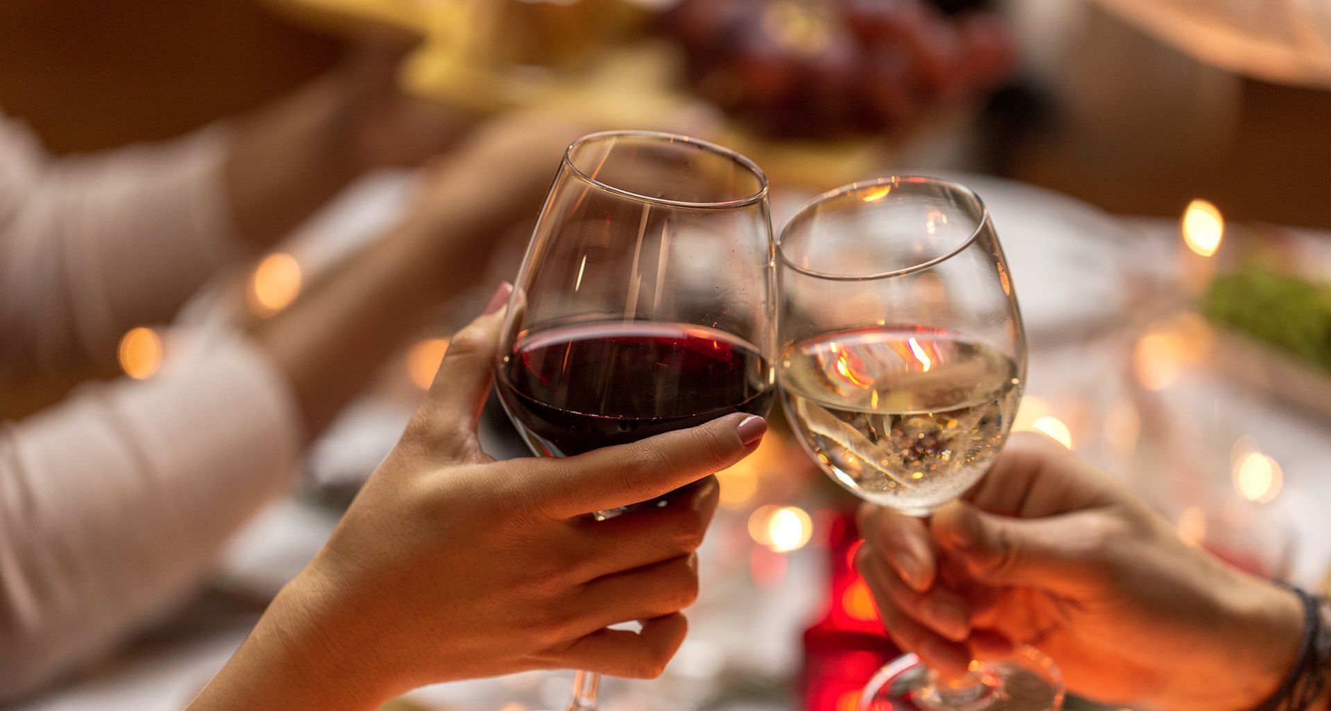 Two hands toast with glasses of red and white wine at a dinner table with out-of-focus lights in the background.