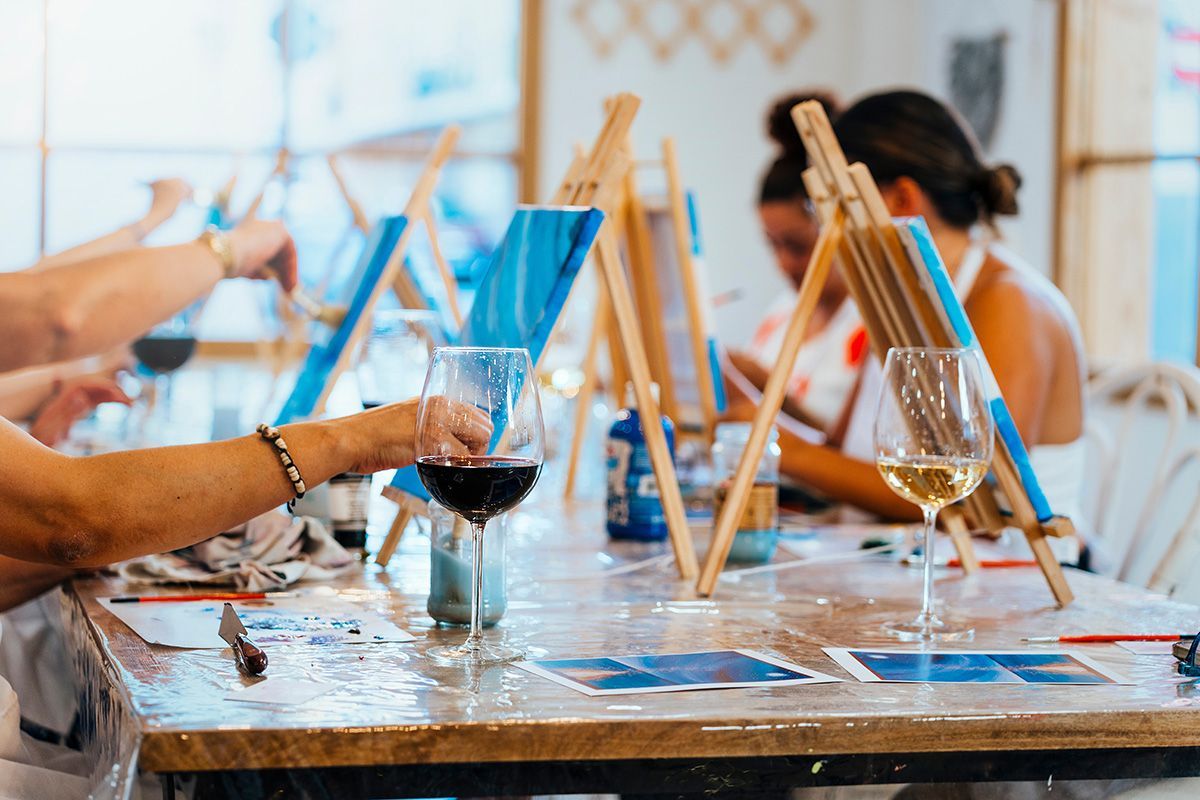 People painting on canvases at a table with wine glasses during an art class.