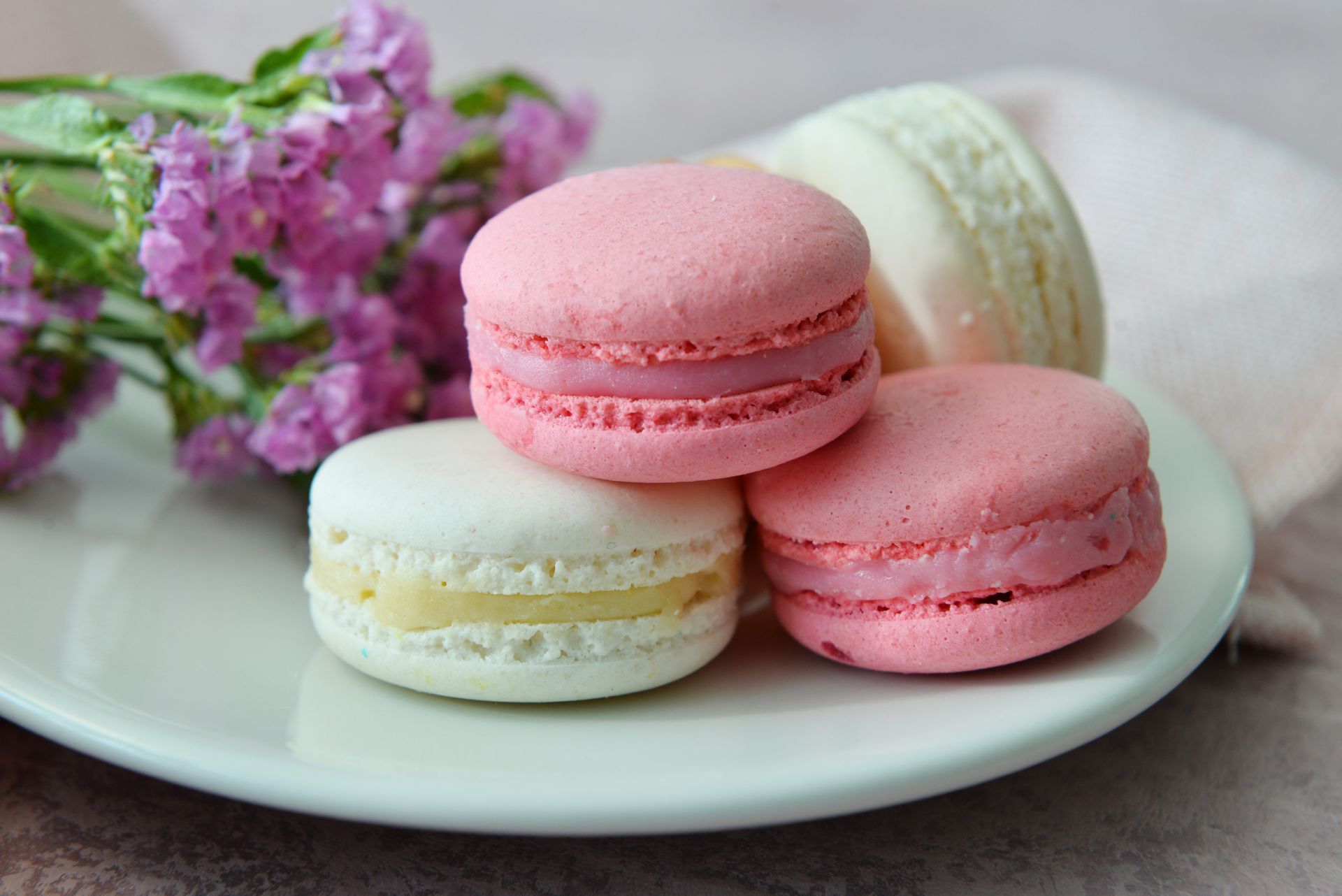 Three macarons in pink and white are arranged on a white plate next to a small bunch of purple flowers.