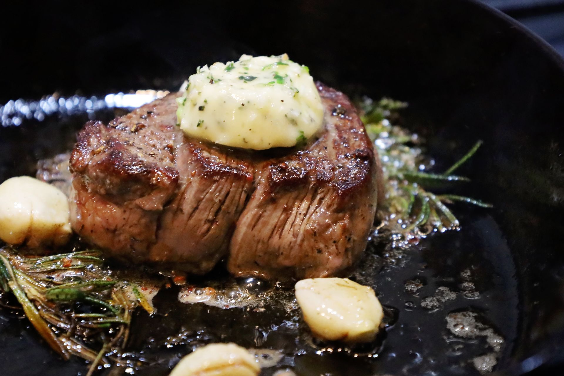 A seared filet mignon in a cast-iron skillet, topped with a disk of herb butter, accompanied by garlic and fresh rosemary.