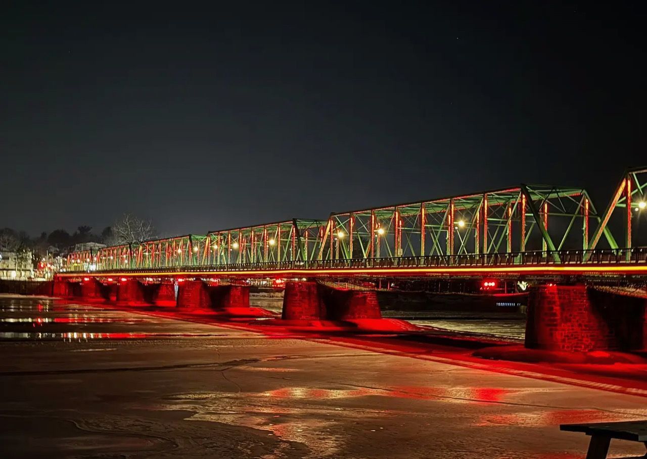 A steel truss bridge at night, illuminated by green upper lights and red lights reflecting on a dry riverbed below.