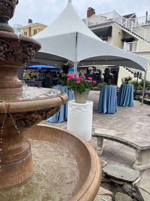 A stone fountain stands in the foreground of a patio set for an event with a white tent and blue-clothed tables.