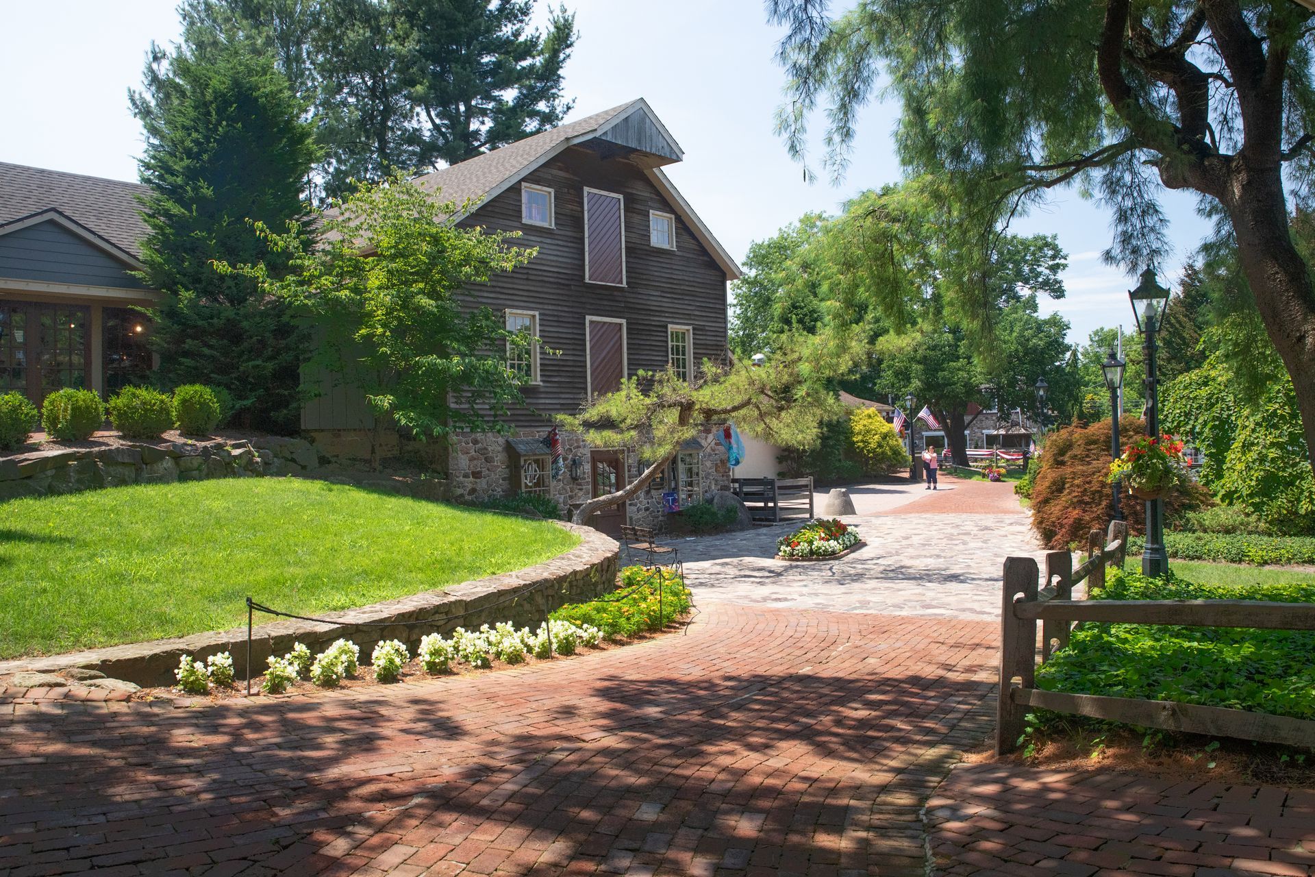 A rustic wooden building with stone foundation sits beside a brick walkway and a green lawn on a sunny day.