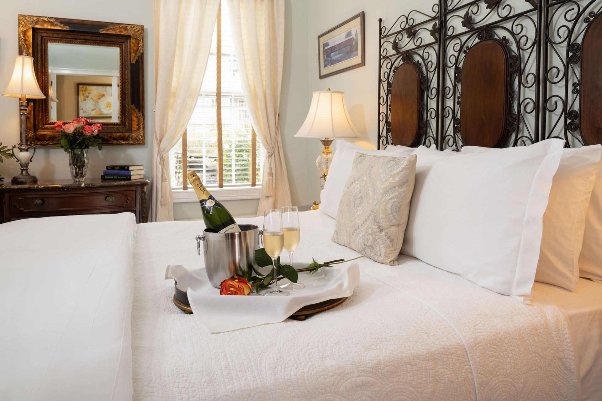 A romantic bedroom setting with white bedding, a champagne bucket, two glasses, and a rose on a tray atop the bed.