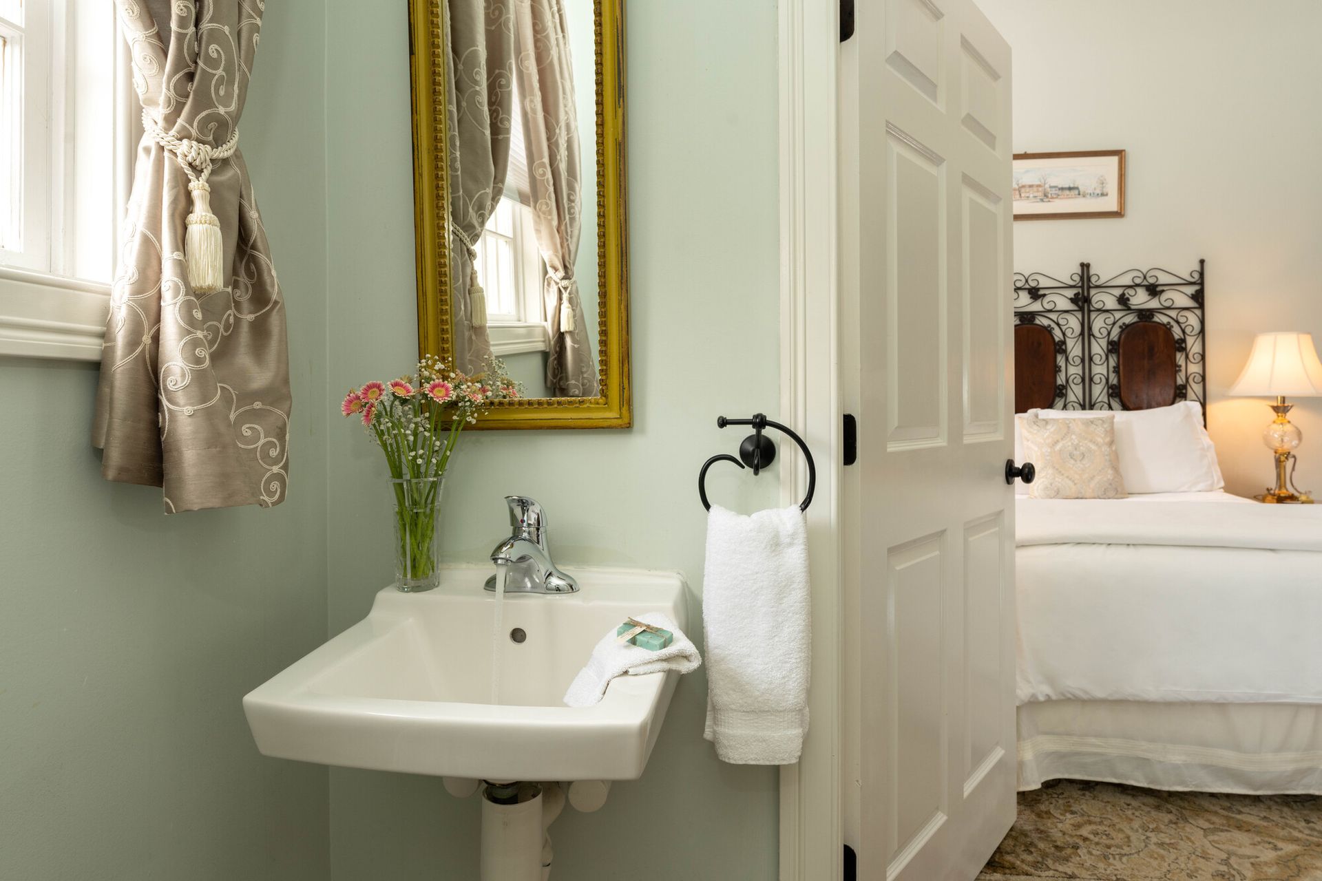 A pedestal sink with flowers next to a doorway showing a glimpse of a bedroom with a white bed and a lamp.