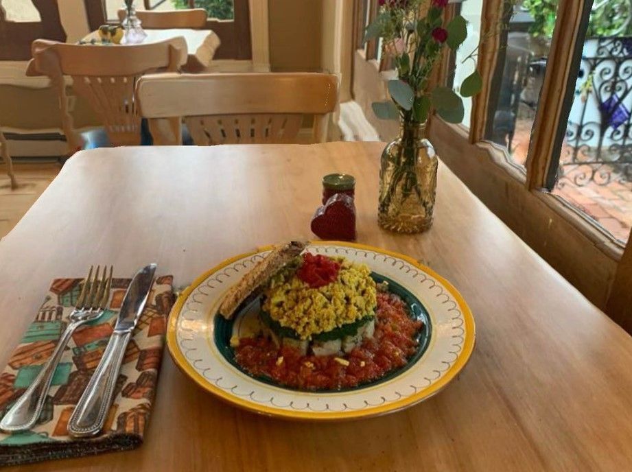 A plate of food featuring yellow salad, tomato salsa, and toast on a wooden table with cutlery and a vase of flowers.