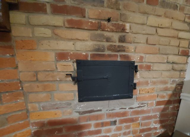 A black rectangular metal oven or clean-out door set into a rustic, multi-toned brick wall.