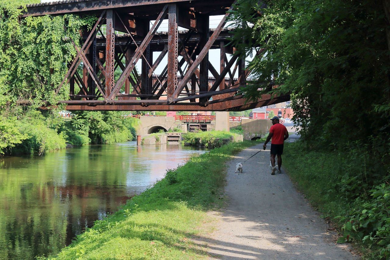 A person in a red shirt walks their small dog along a gravel path beside a canal, under an old rusted metal bridge.