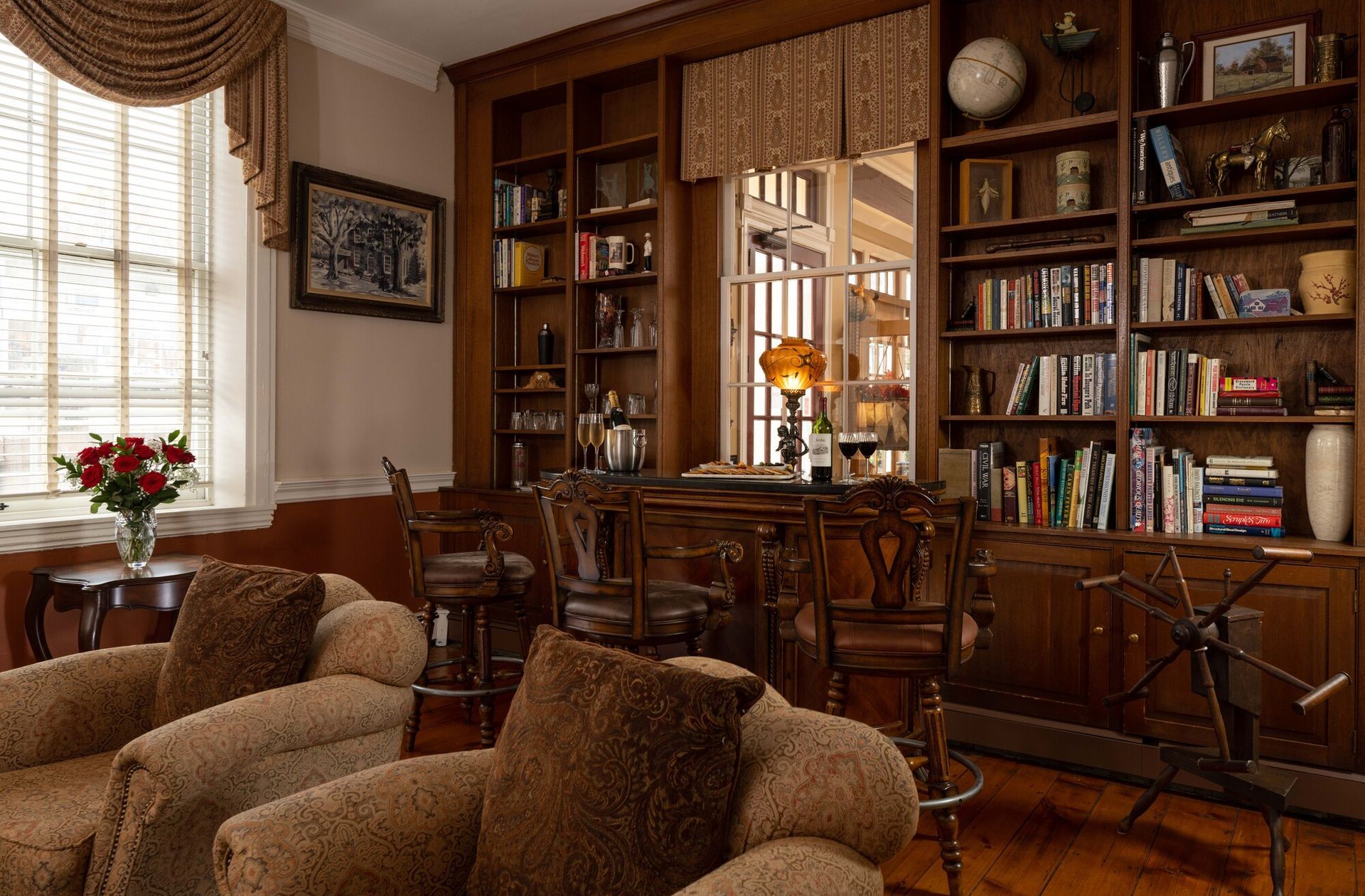A cozy library with plush chairs, floor-to-ceiling wooden bookshelves, and a window with curtains.