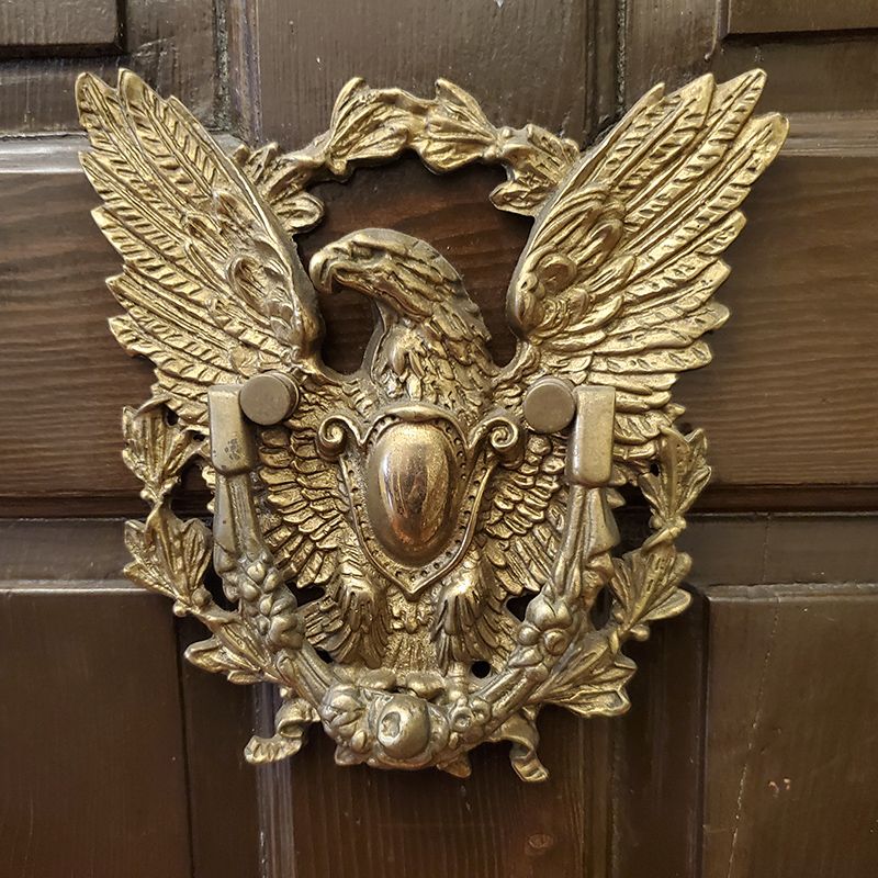 A brass eagle door knocker mounted on a dark wooden door, featuring an eagle with wings spread above a central shield.