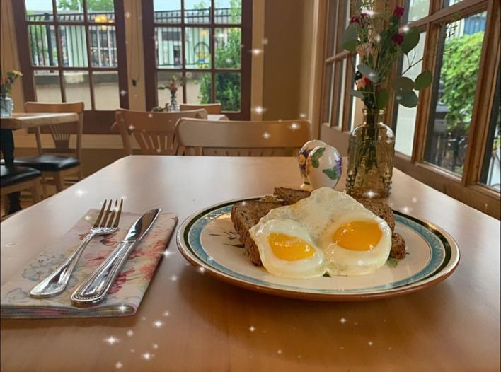 A plate with two sunny-side-up eggs on toast sits on a wooden table beside cutlery and a vase of flowers in a cafe.