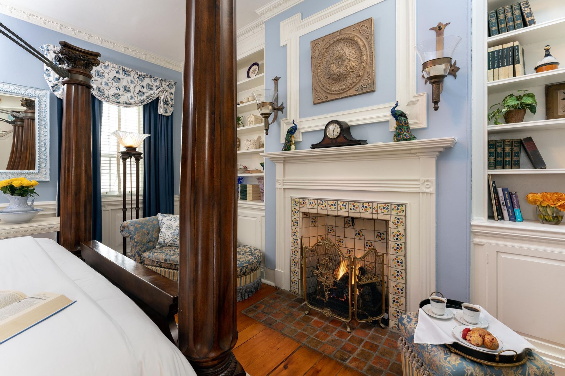 A blue-walled bedroom with a four-poster wooden bed, fireplace, bookshelves, and a small chair by the window.