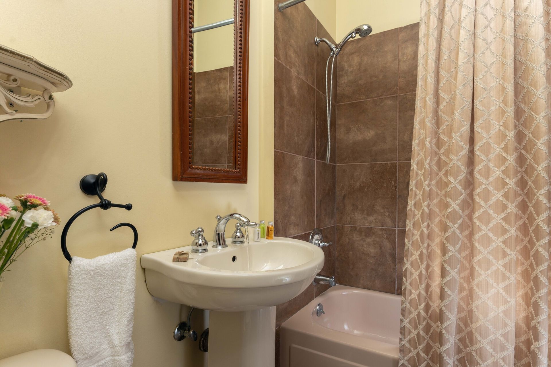A bathroom with a pedestal sink, a wood-framed mirror, a beige towel on a black ring, and a tub with brown tiled walls.