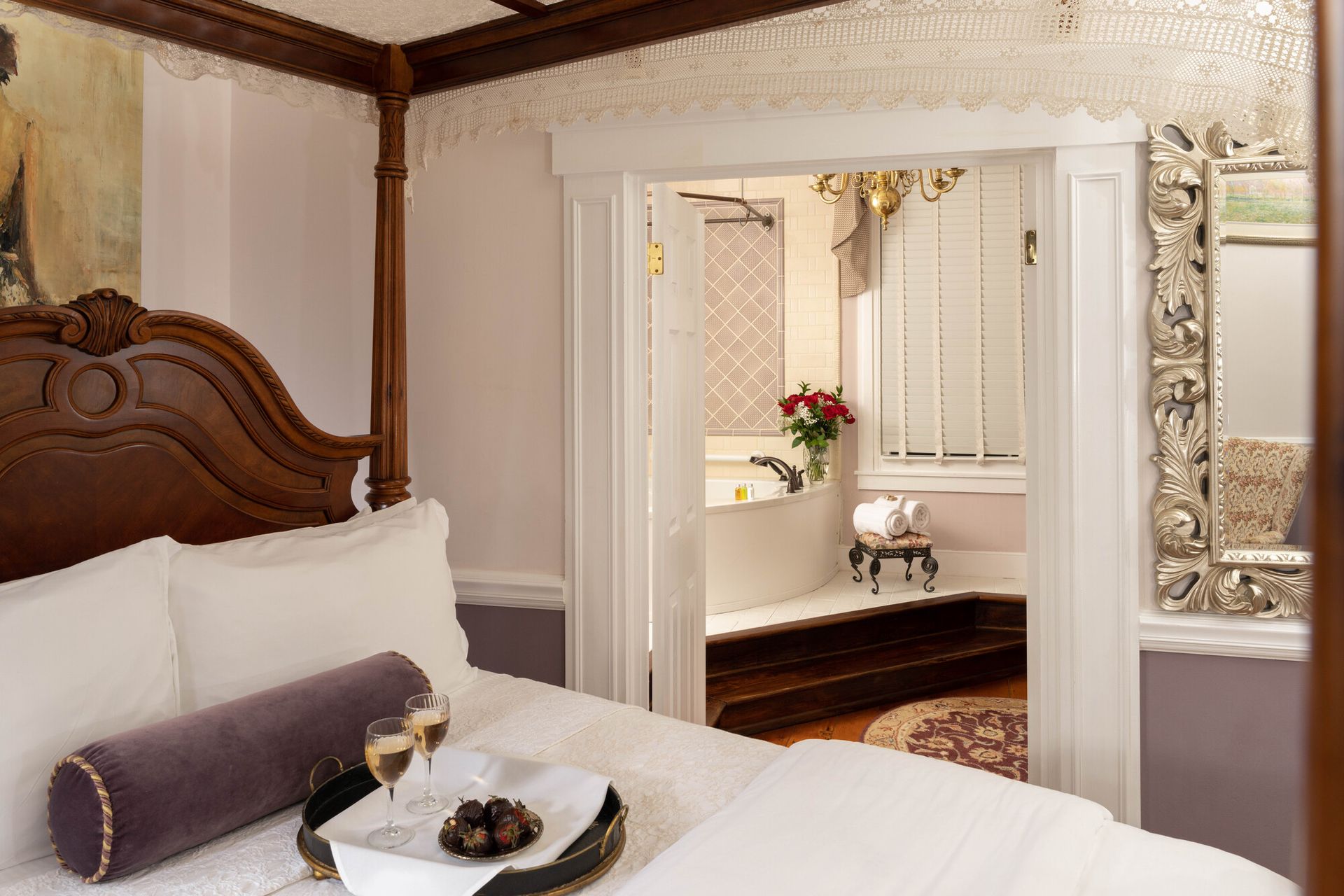 A cozy bedroom with a four-poster bed, a tray with drinks, and an open doorway leading to a bright, elegant bathroom.