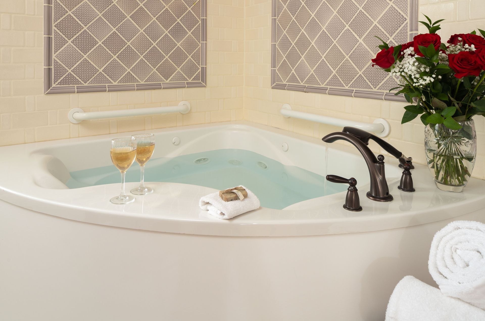A corner bathtub filled with water, two glasses of wine, a folded towel, and a vase of roses on the ledge.