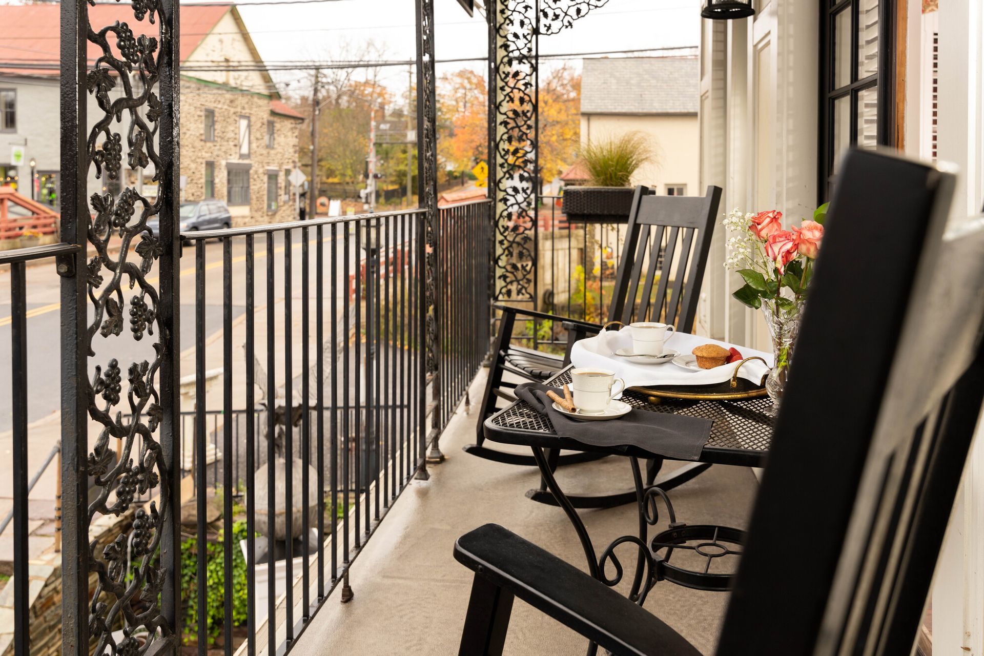 Two black rocking chairs sit on a balcony with a small table featuring tea and flowers, overlooking a street.