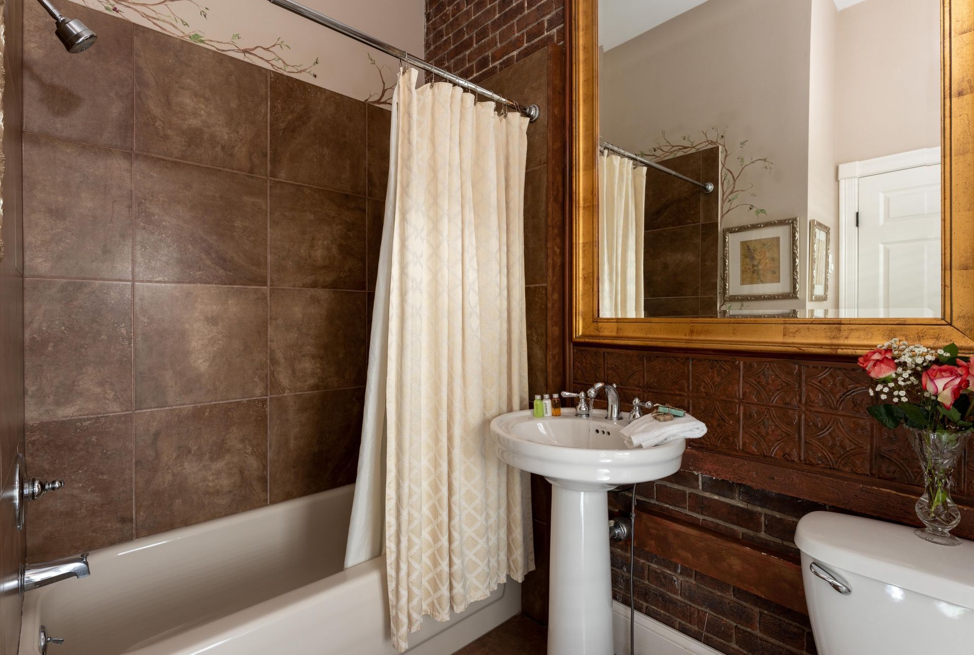 A bathroom with a brown tiled shower, a white pedestal sink, a toilet, and a mirror in a wood frame against a brick wall.