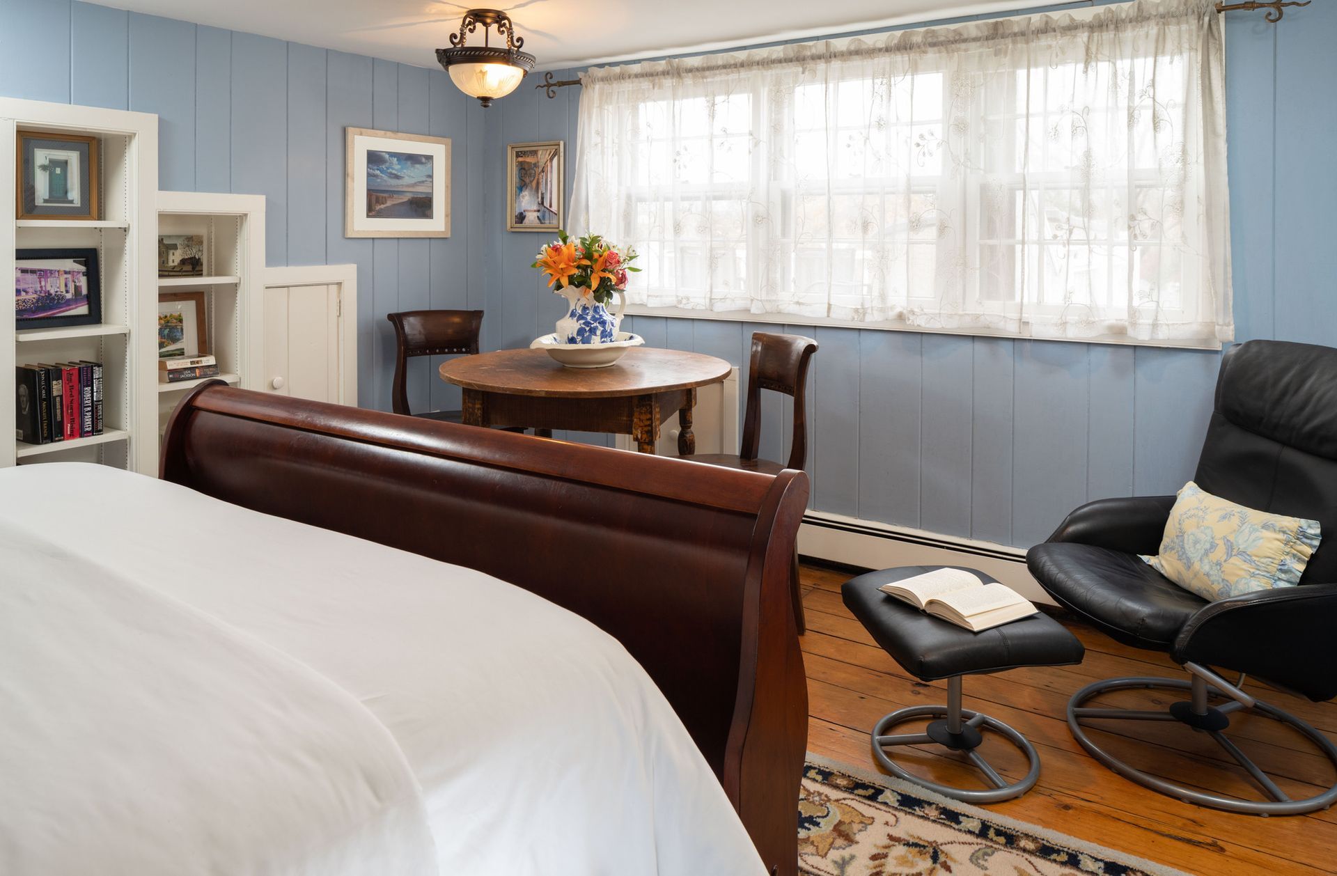 A bedroom with a dark wood bed, a small round table with flowers, and a black leather armchair with an ottoman.