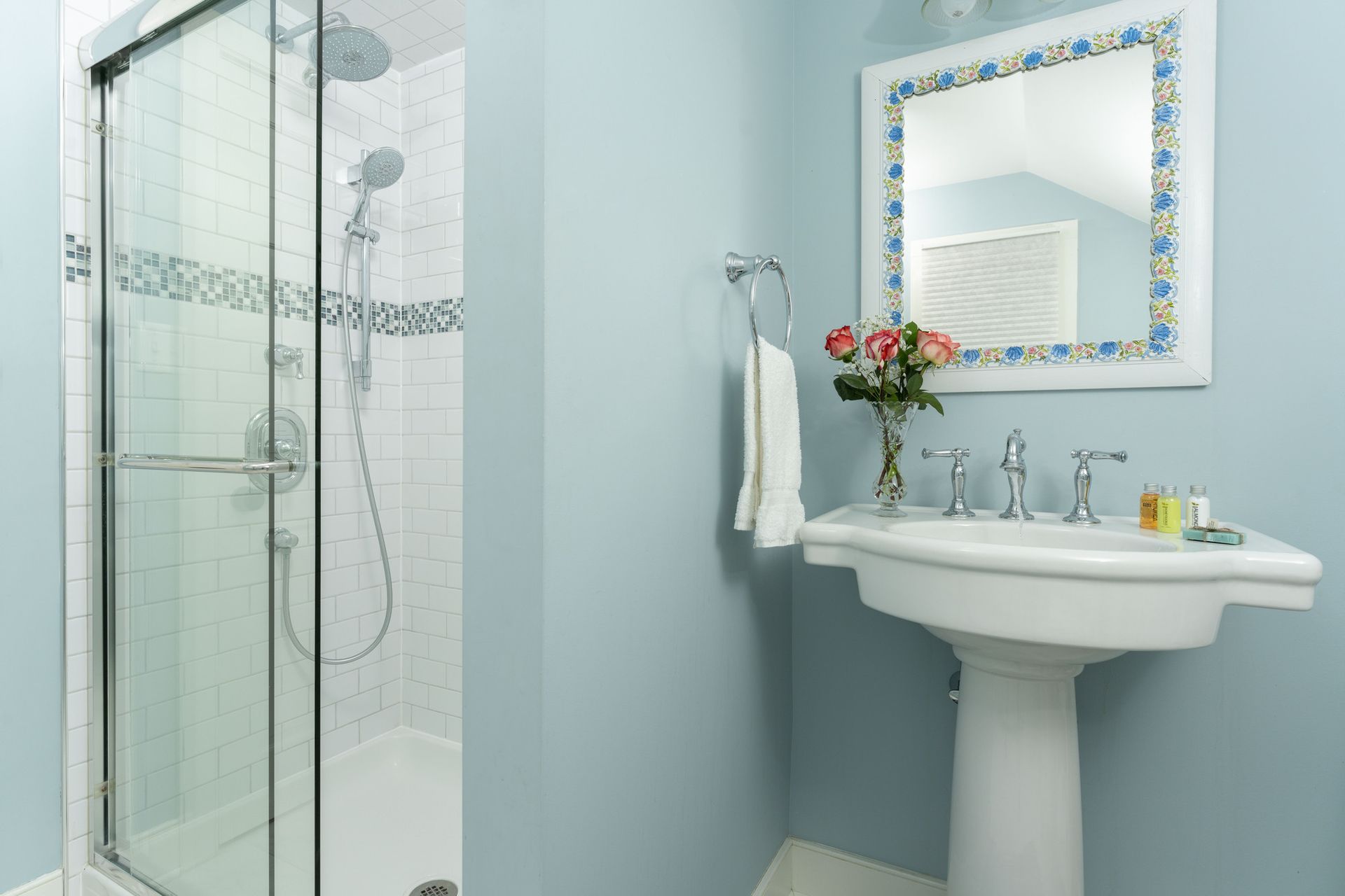 A light blue bathroom with a glass-enclosed shower, a pedestal sink, and a mirror with a blue and white patterned frame.