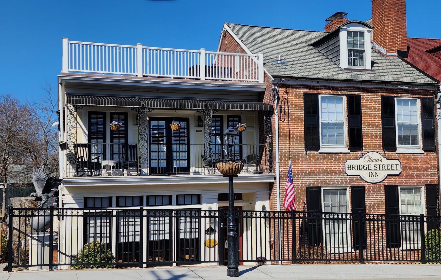 A white multi-story house with a balcony and rooftop deck sits beside a brick building with black shutters and a sign.