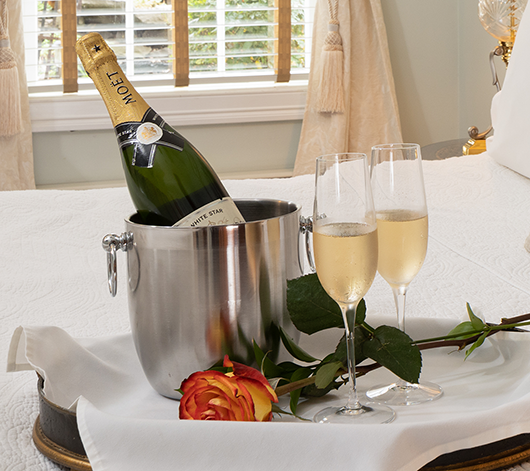 A champagne bottle in a silver bucket, two filled glasses, and a rose on a white tray set on a bed.