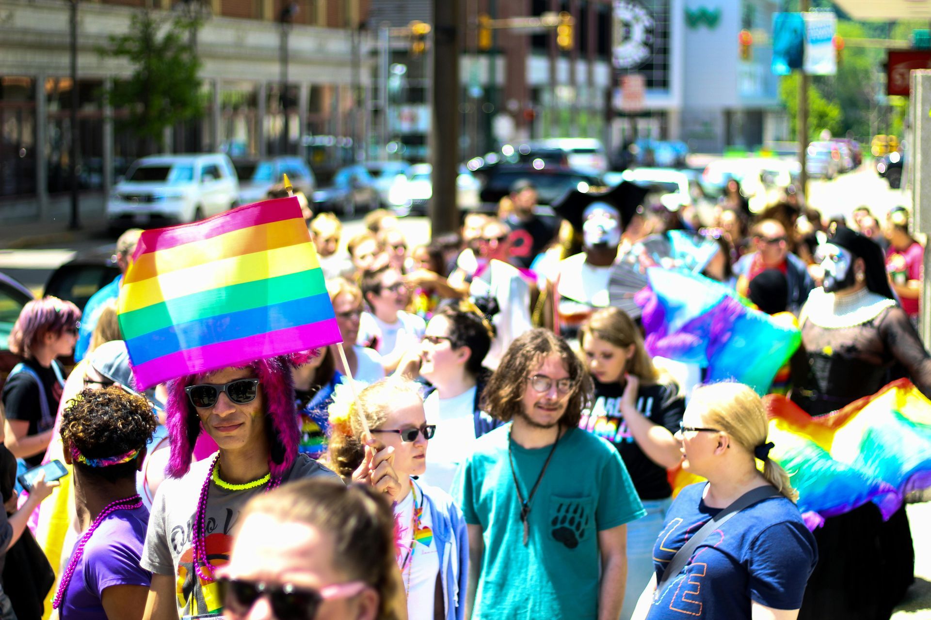 A crowd walks down a city street during a Pride parade, featuring bright rainbow flags and celebratory clothing.
