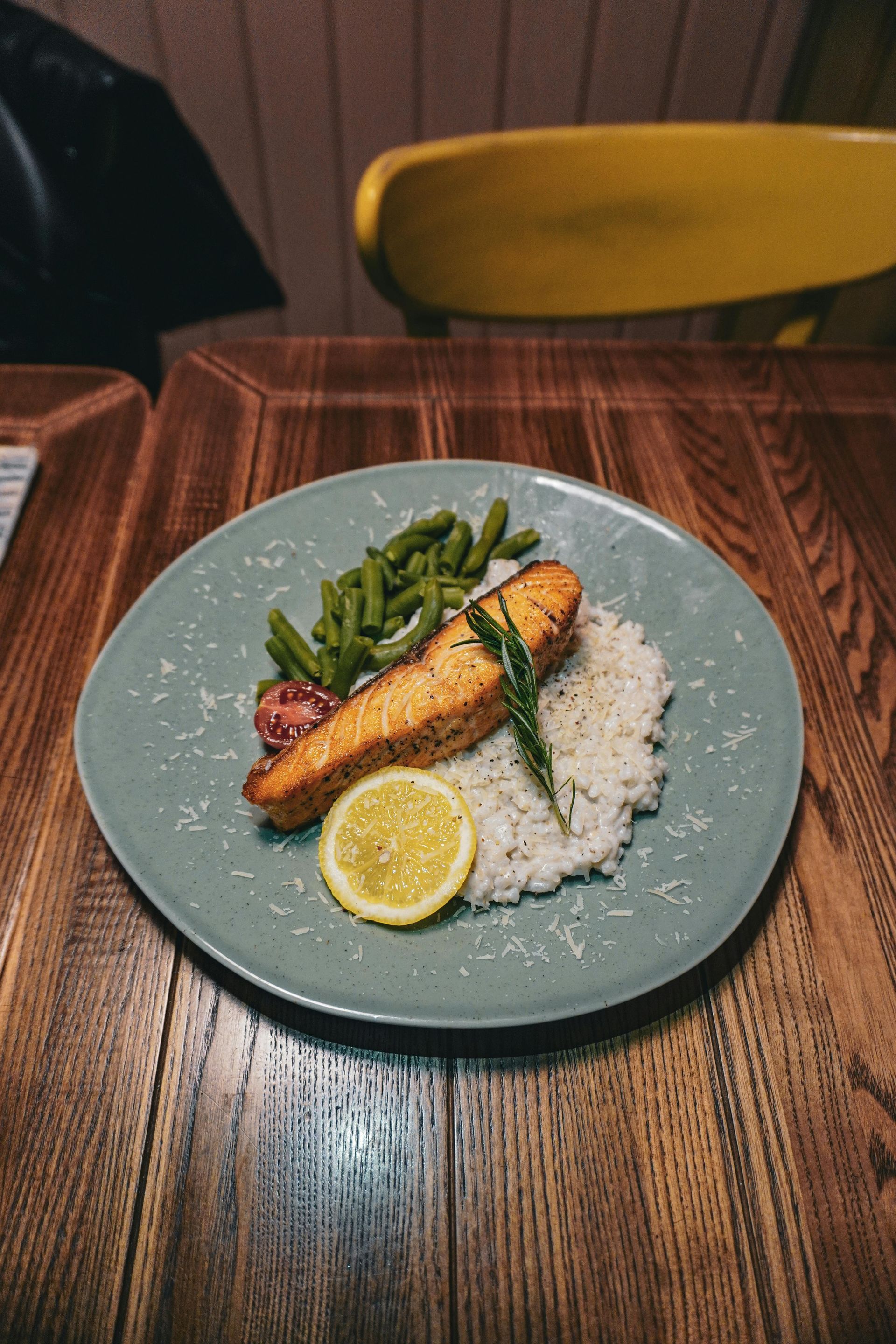 A salmon fillet served with white rice, green beans, and a lemon slice on a blue plate atop a wooden table.