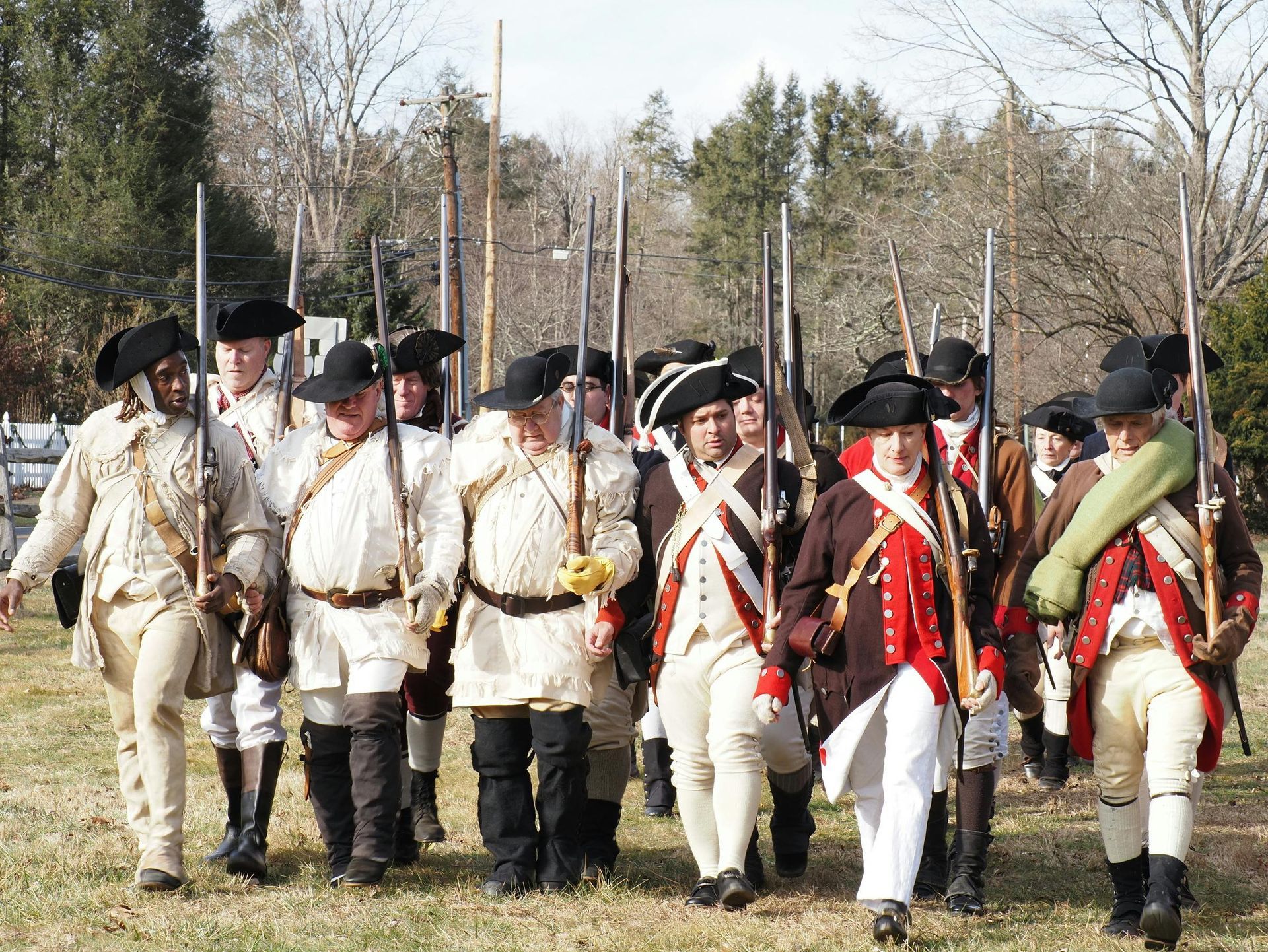 Reenactors in 18th-century colonial military uniforms march in a field, carrying muskets during an outdoor event.