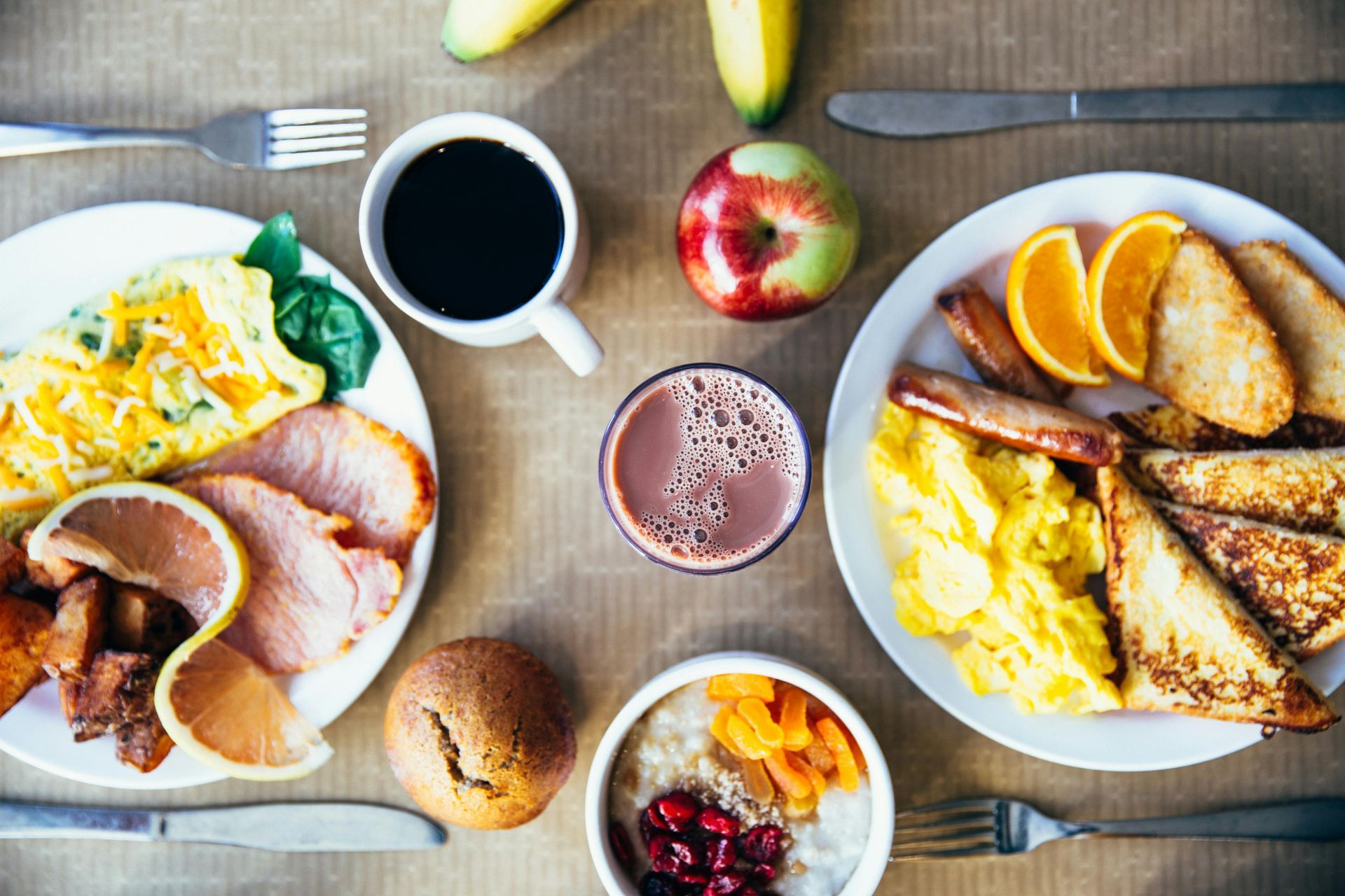 Top-down view of a breakfast spread with two plates of eggs, meat, toast, fruit, a bowl of oatmeal, and drinks.