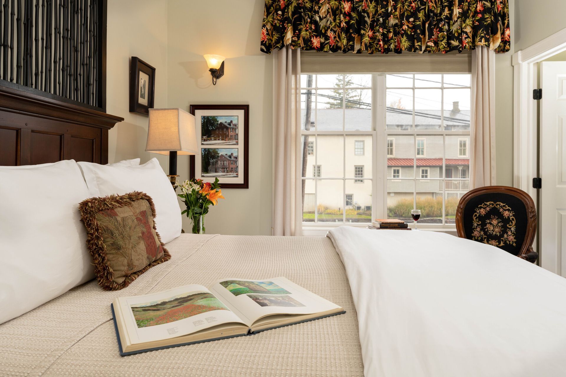 An open book lies on a bed with white linens and a decorative pillow in a room with a large window and floral valance.
