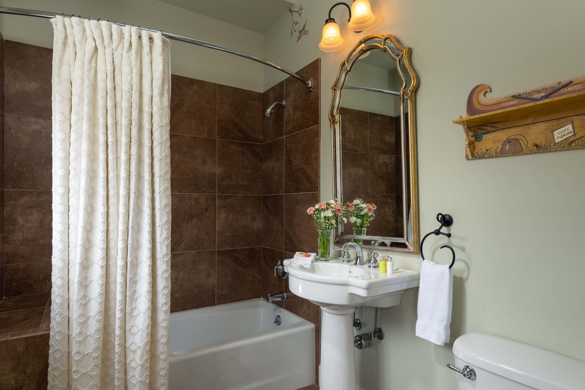 A bathroom with a white pedestal sink, a bathtub with a white ruffled shower curtain, a gold-framed mirror, and a shelf.