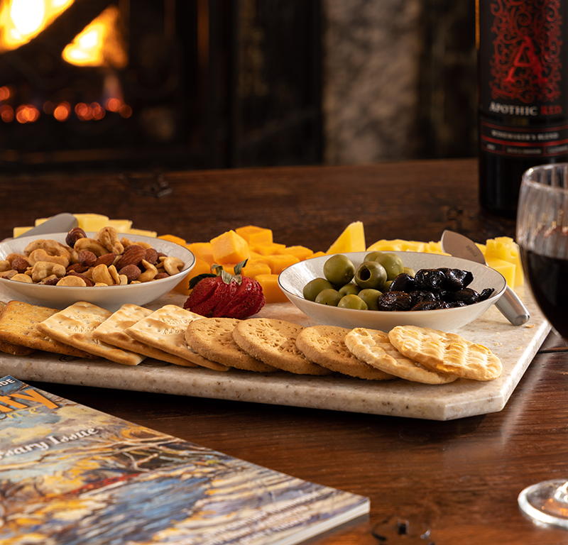 A charcuterie board with crackers, nuts, olives, and cheese sits on a wooden table before a fireplace, with red wine nearby.