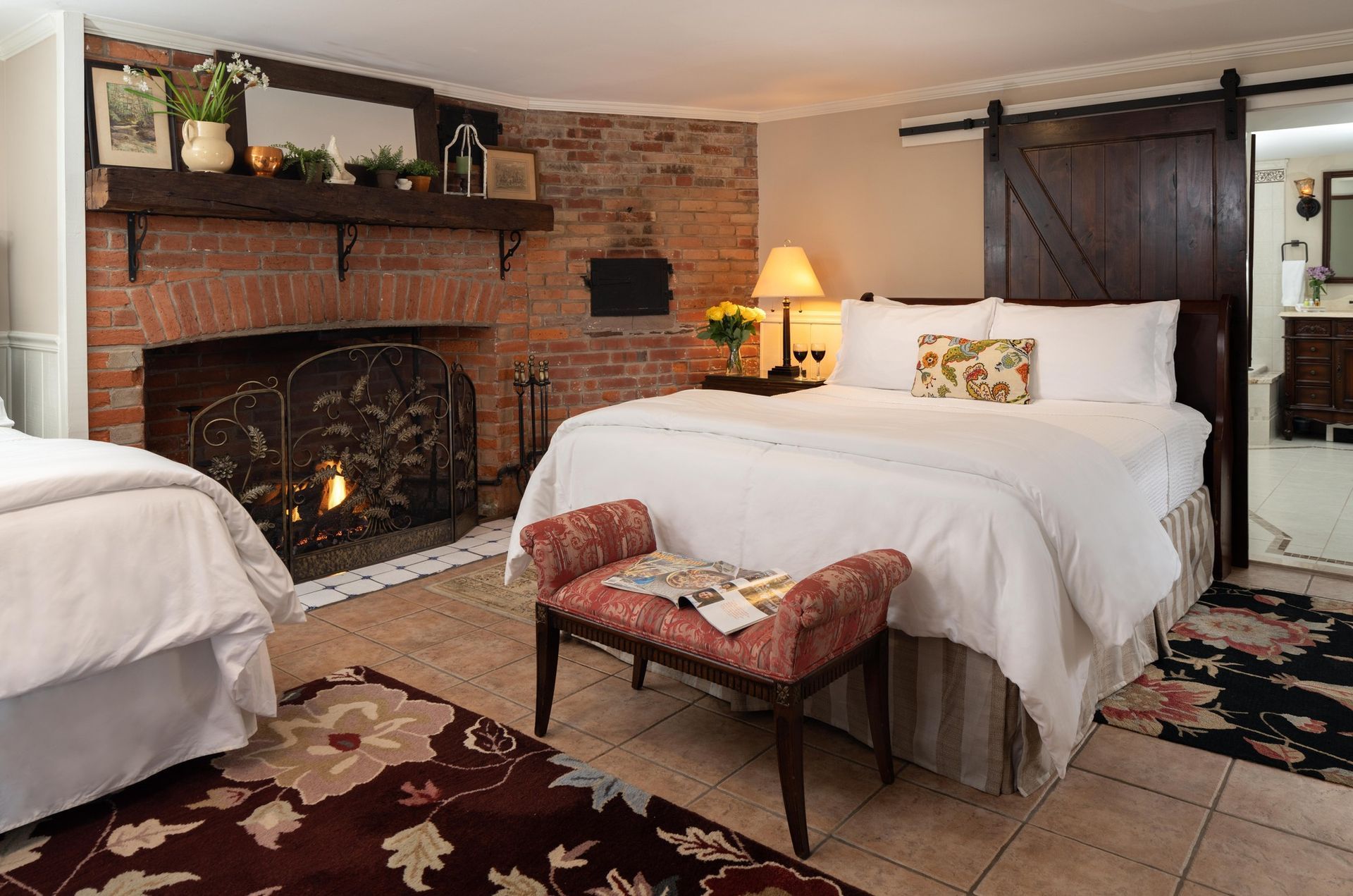 A hotel room with a fireplace, a queen-sized bed, a patterned bench, and a dark sliding door leading to a bathroom.