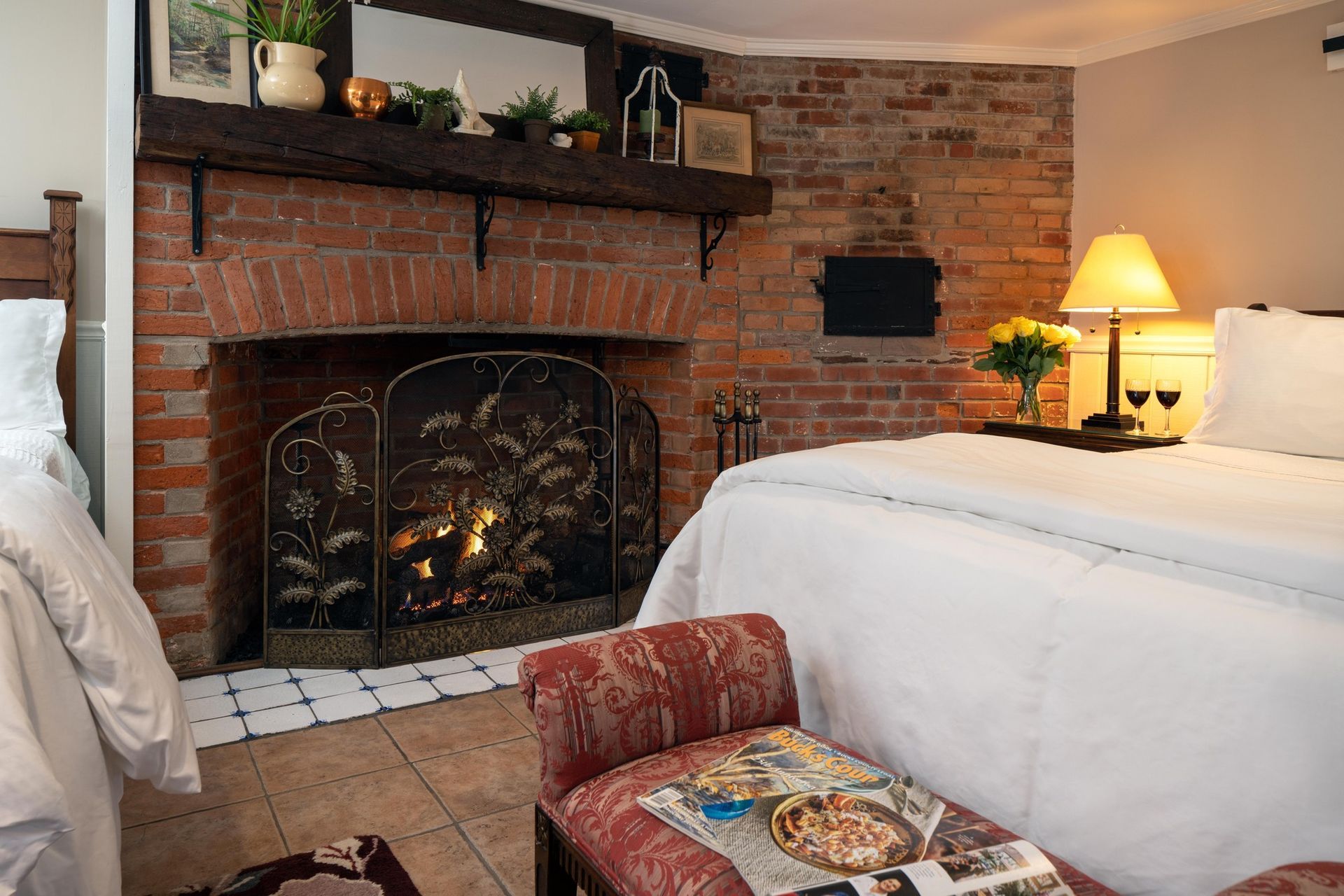 A cozy hotel room featuring a brick fireplace with a decorative metal screen, a plush bed, and a footstool with a tray.
