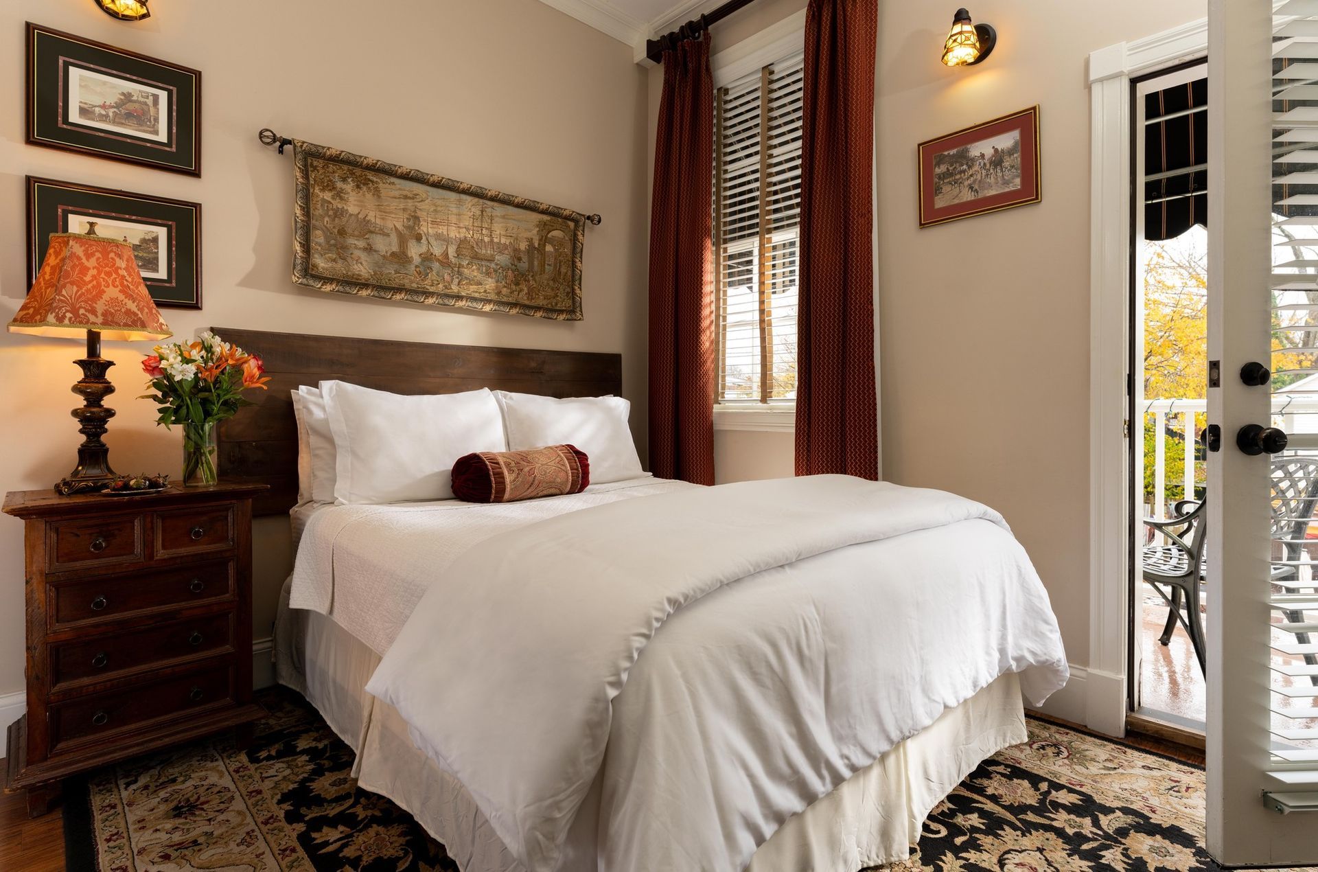 A cozy bedroom with a white-made bed, dark wood nightstand with a lamp, decorative wall art, and an outdoor balcony door.