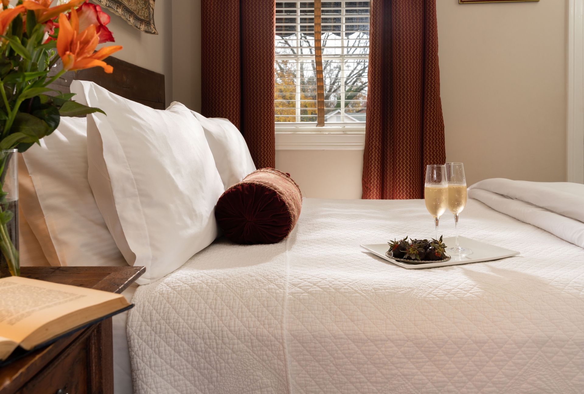 A hotel bed with white linens, pillows, a decorative bolster, a book on a nightstand, and two glasses of champagne.