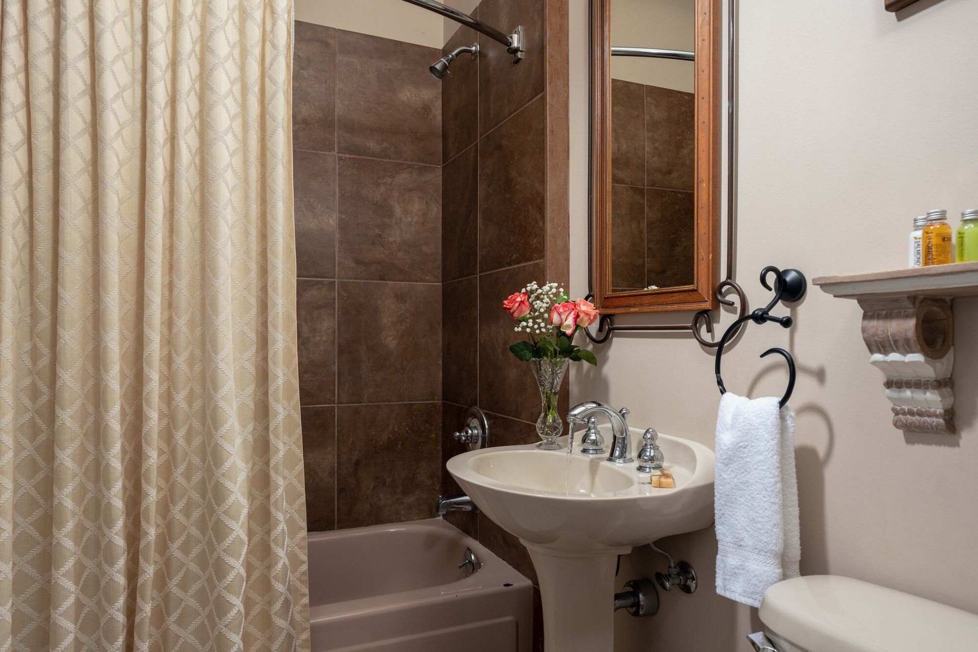 A bathroom featuring a beige shower curtain, a pedestal sink with flowers, a mirror, and a toilet with toiletries nearby.