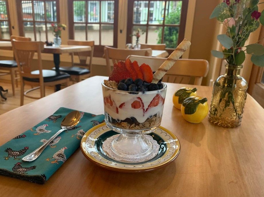 A fruit yogurt parfait with a wafer stick in a glass on a saucer, beside a spoon and vase on a wooden cafe table.