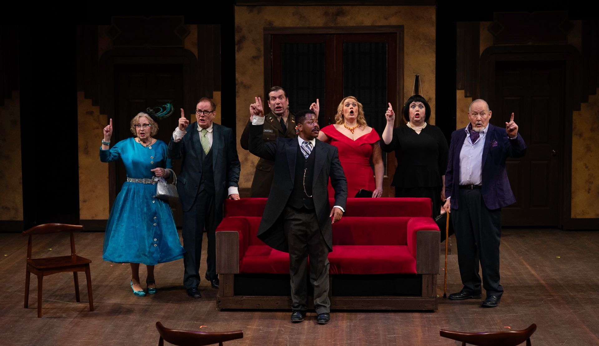 Group of actors on stage, pointing up, dressed in formal attire. Red couch in the center. Dark background.
