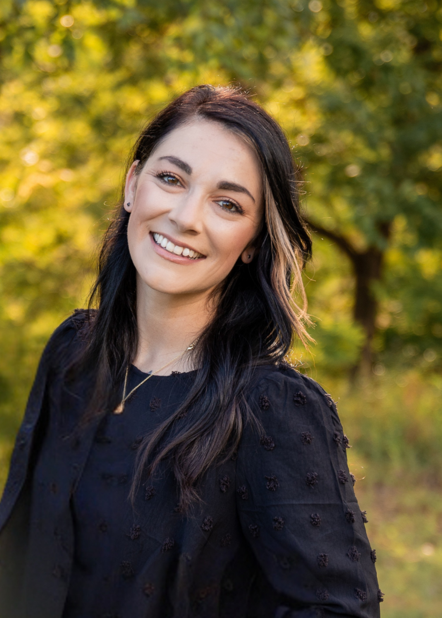 Woman smiling at the camera, dark hair with blonde highlights, wearing a black shirt, set outdoors.