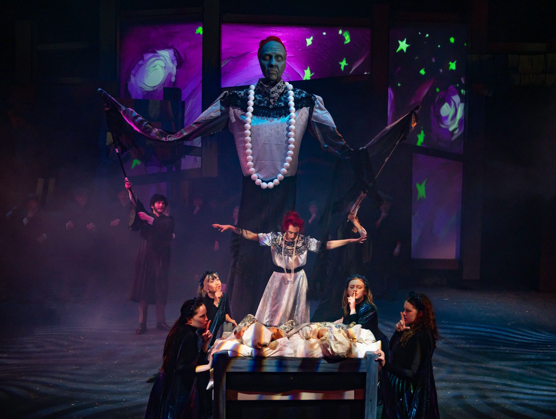 Stage production with a large figure above a person with outstretched arms, surrounded by others, dark setting.