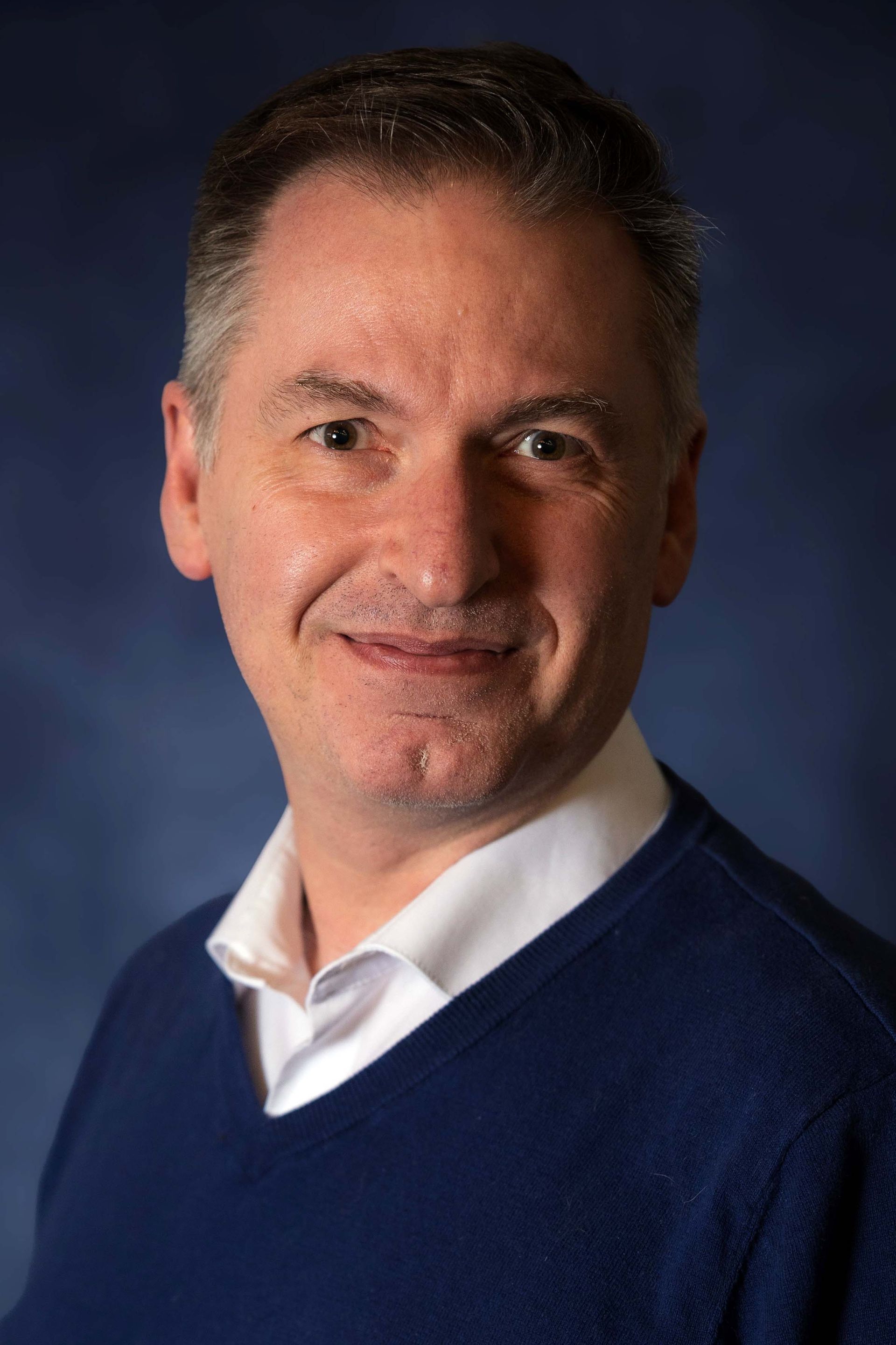 Man with short graying hair, smiling, wearing a blue sweater over a white collared shirt, blue background.