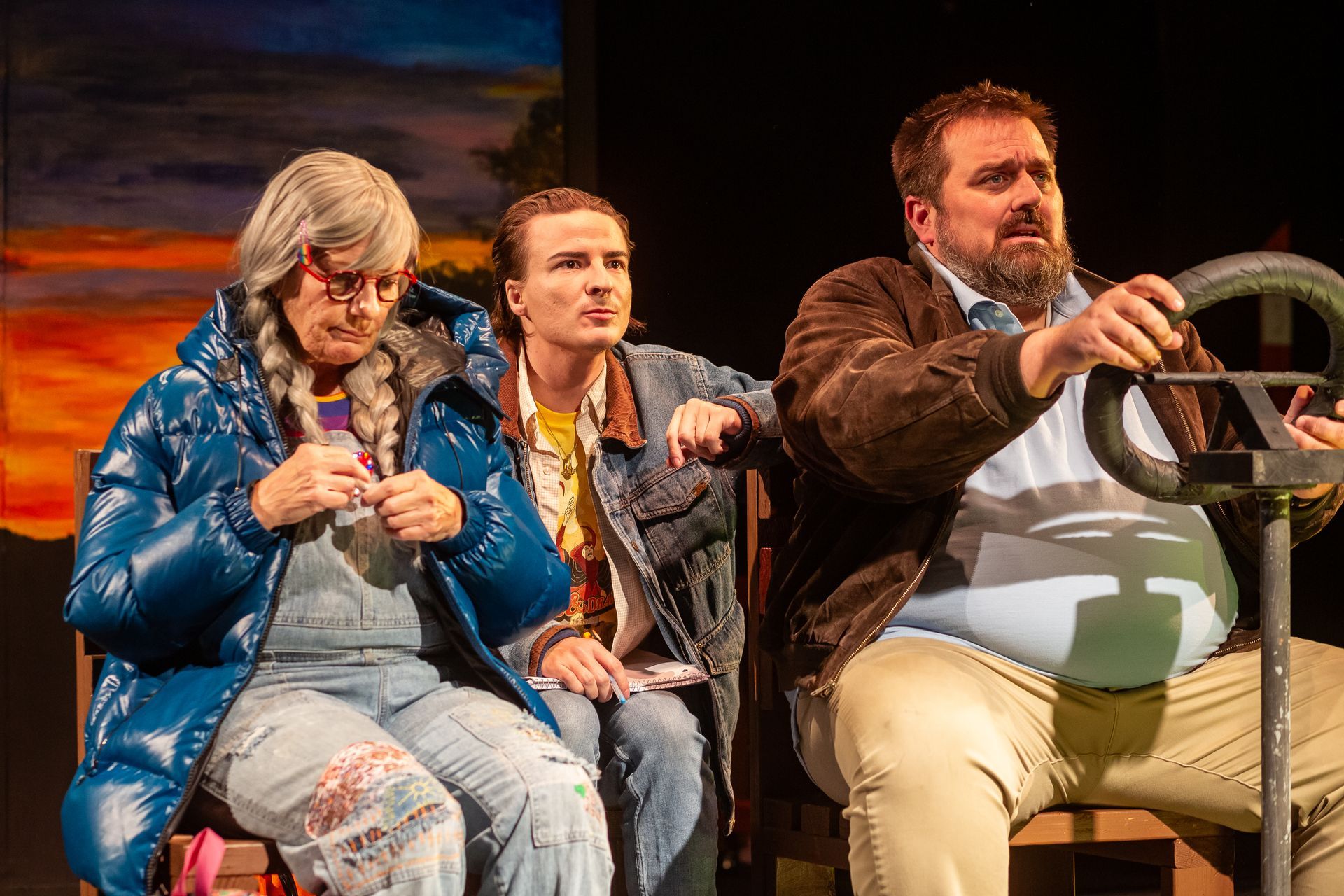 Three people sit on a bench in a theatrical set. One wears a blue coat, one a denim jacket, and one holds a steering wheel.