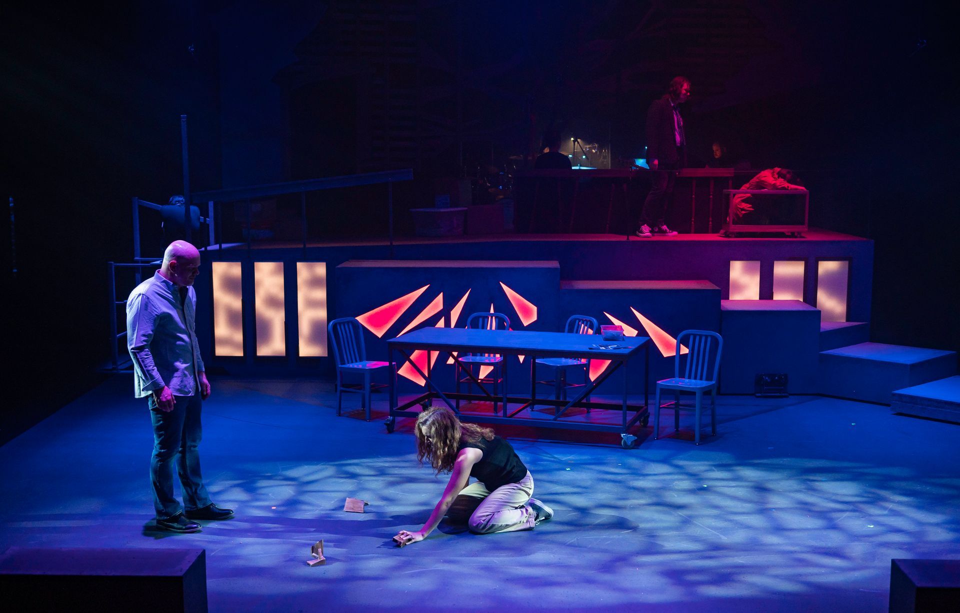 Stage with two figures, a man and a woman, under blue and red lights. Woman kneels, man stands.