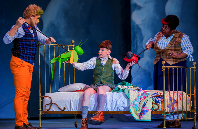 Three people on stage, one on a bed holding objects; others holding puppet strings.