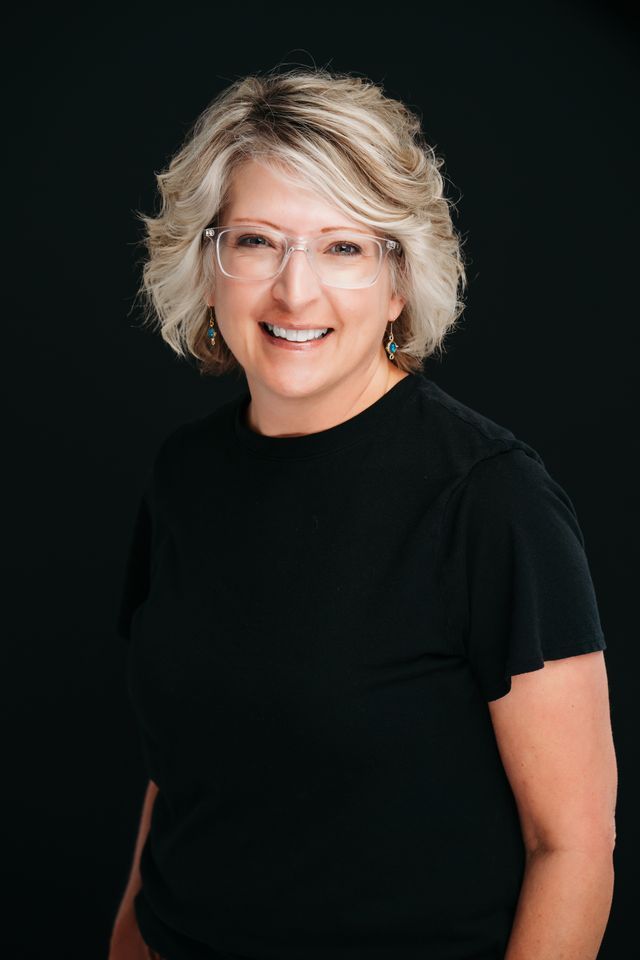 Woman with glasses smiling, wearing a black shirt, against a dark background.