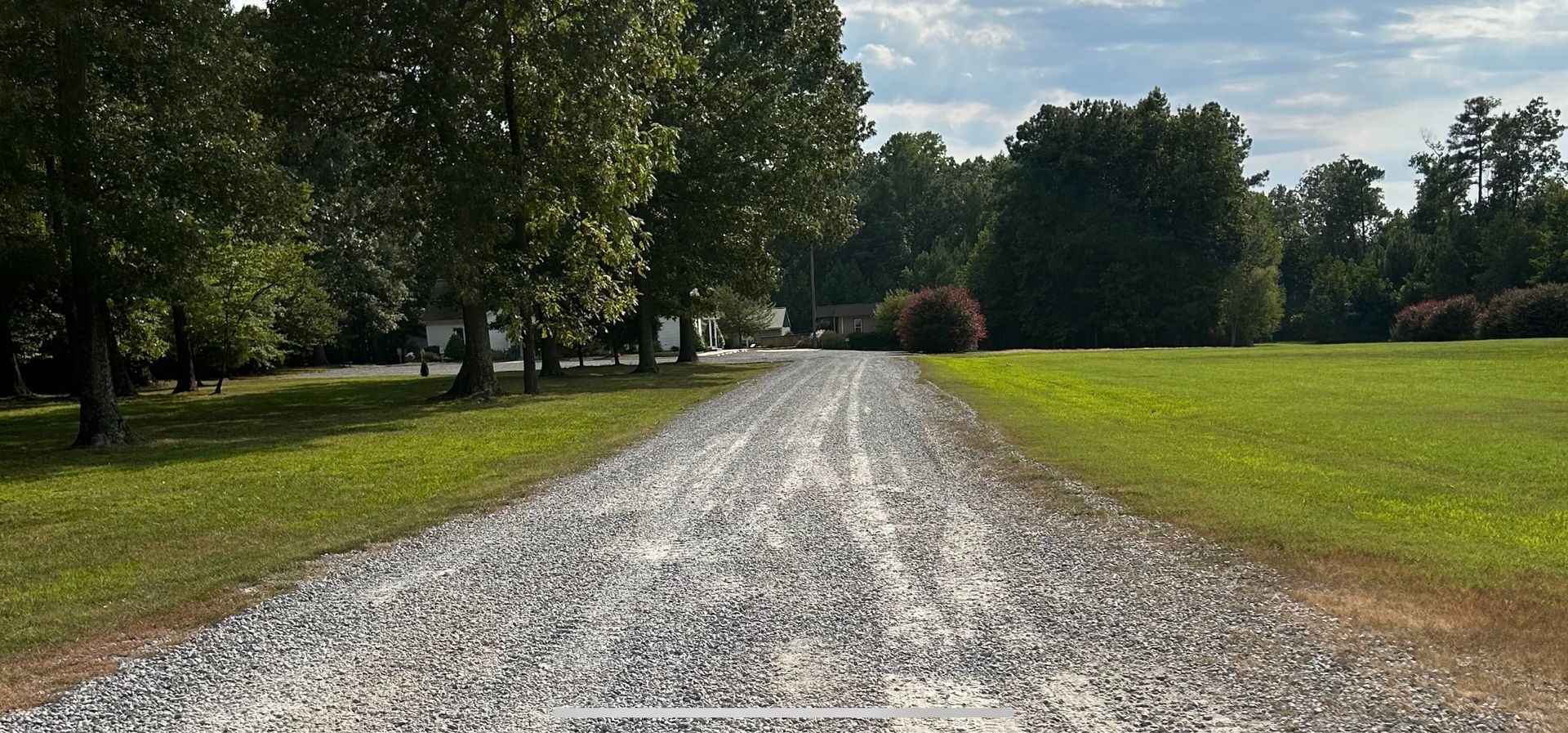 Gravel driveway leading toward trees and a building, with grassy areas on both sides. Cloudy sky.