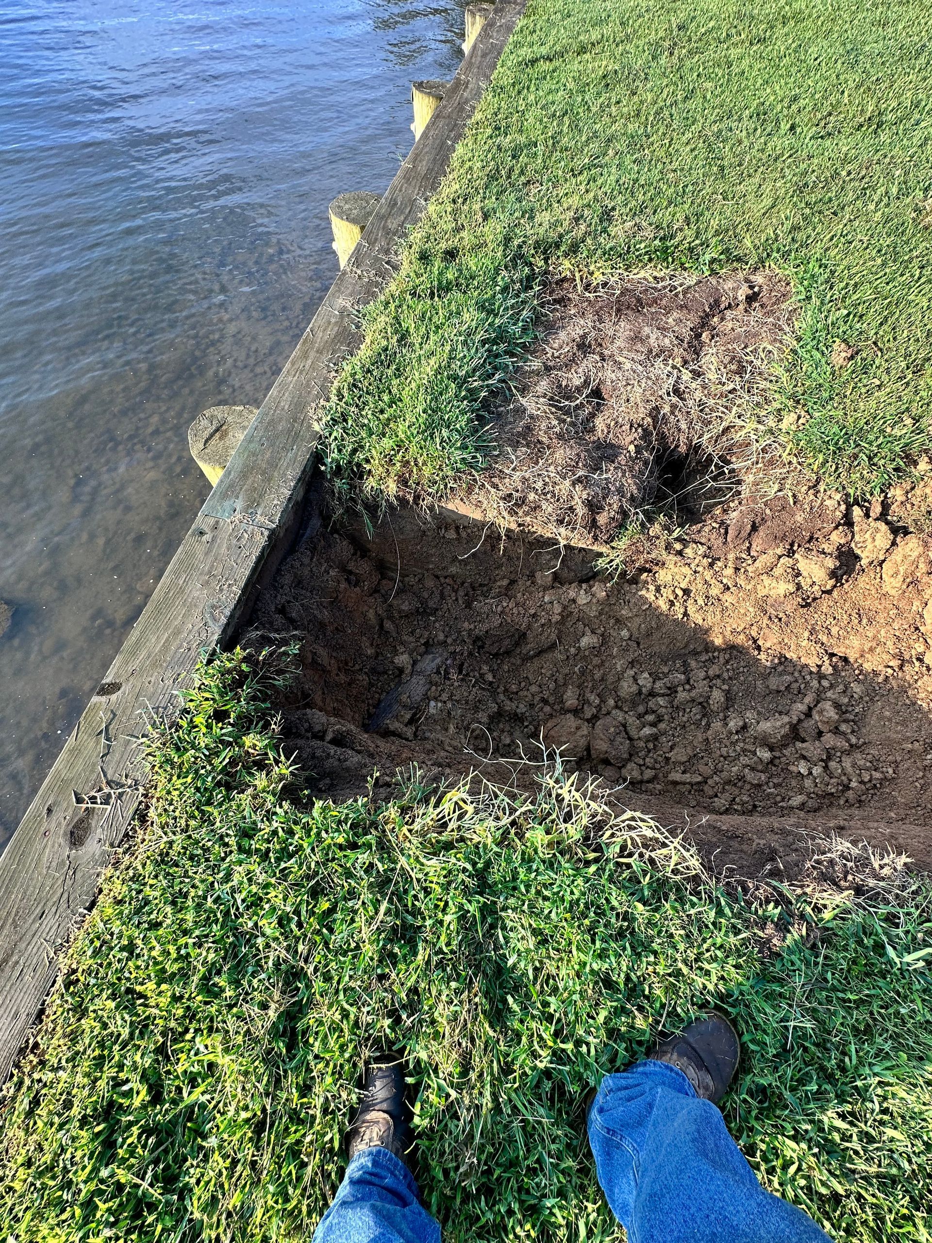 Erosion damage next to water. Dirt and missing grass. Person's feet wearing boots at bottom of frame.