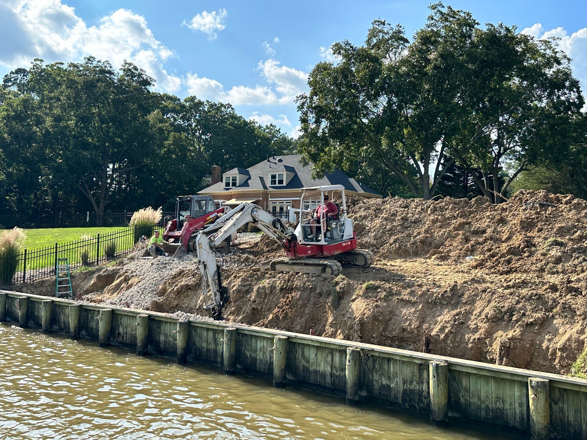 Excavators removing earth next to a house on a waterway. Wooden bulkhead in the foreground, trees in the background.