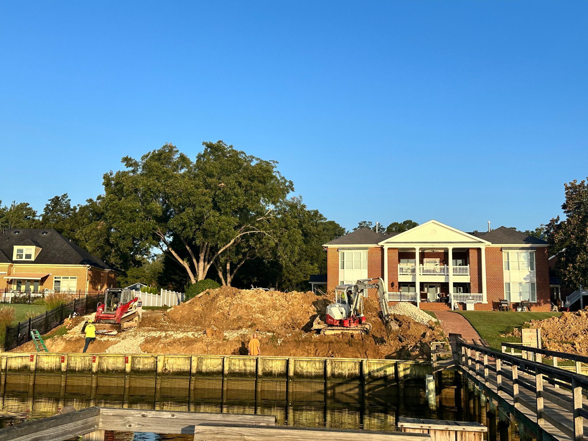 Construction site next to a lake with excavators, a large pile of dirt, and a multi-story brick house.